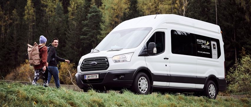 Ein Mann und eine Frau gehen neben einem weißen Ford Transit-Van mit einer Postbus-Shuttle-Werbung an der Seite, vor einem Wald.