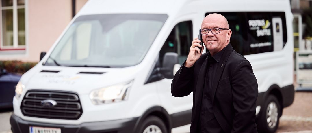 Ein kahlköpfiger Mann in einem schwarzen Anzug spricht auf einem Handy, während er vor einem weißen Lieferwagen steht.