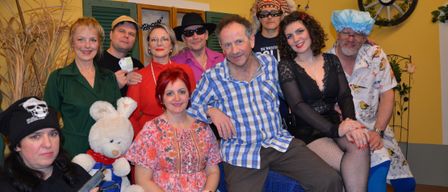 A group of people in various costumes pose for a photo. A man in a plaid shirt sits next to a woman in a black dress. A white teddy bear and a toy gun are present.