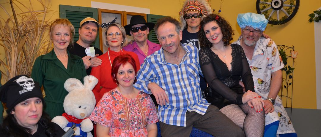 Eine Gruppe von Menschen in Kostümen posiert für ein Foto, einer hält ein Plüschtier. Ein Mann in einem blau-weiß karierten Hemd sitzt auf einem Stuhl.