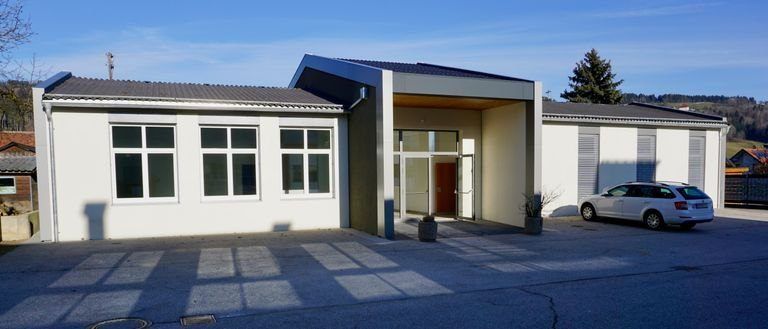 Ein weißes Gebäude mit grauem Dach und Glasfenstern ist zu sehen. Ein Auto ist vor dem Gebäude geparkt. Es gibt zwei Topfpflanzen an der Seite. Bäume und Häuser sind in der Ferne sichtbar.