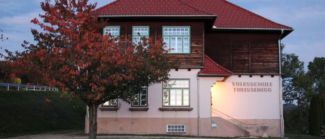 Ein Gebäude mit rotem Dach und weißen Wänden wird vom untergehenden Sonnenlicht beleuchtet. Das Gebäude hat einen Baum davor und mehrere Fenster. Das Schild steht für Volksschul Theissenegg.