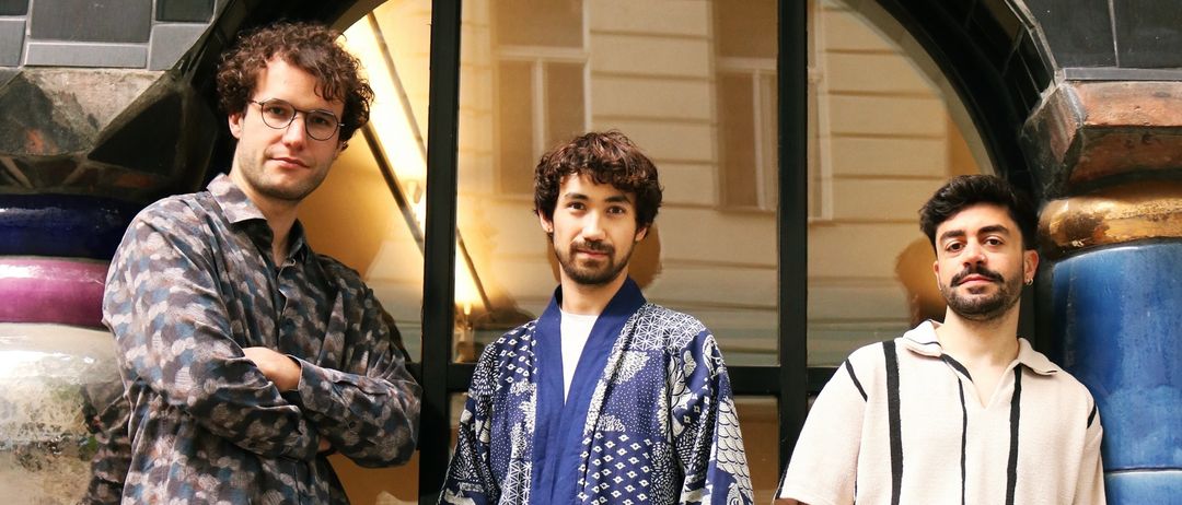 Three men stand together in front of a building. The middle man wears a blue and white patterned robe. The man on the left has curly hair and wears glasses. The man on the right has a beard and wears a striped shirt.