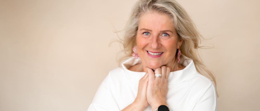A woman with blonde hair is wearing a white shirt, pink earrings, and a watch on her wrist. She is smiling and posing for a photo.
