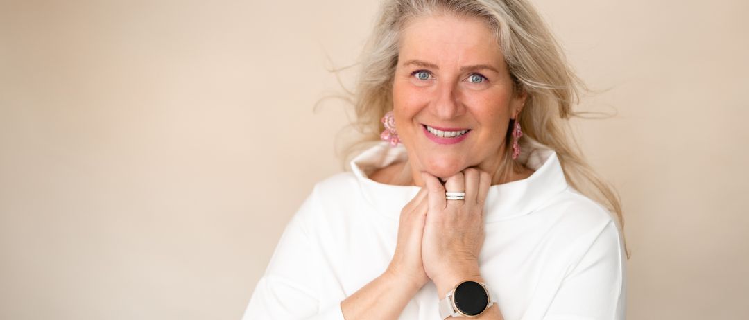 Eine Frau mit grauen Haaren lächelt und posiert für ein Foto. Sie trägt ein weißes Hemd, rosa Ohrringe, eine silberne Uhr und einen Ring. Ihr Haar weht im Wind.