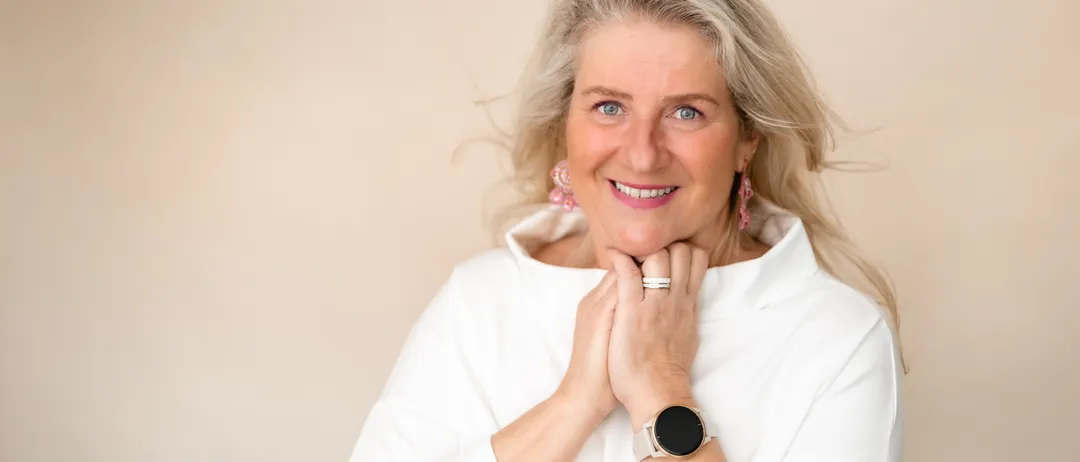 Eine lächelnde ältere Frau mit blonden Haaren posiert für ein Foto. Sie trägt ein weißes Hemd, rosa Ohrringe und eine Uhr. Sie hat ihre Hände vor sich verschränkt.