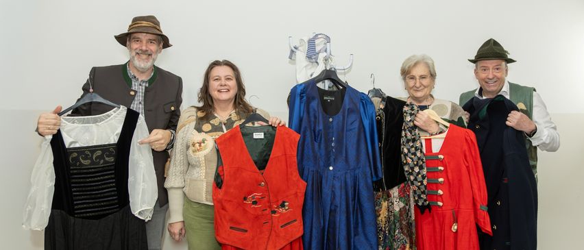 Eine Gruppe von Menschen posiert mit verschiedenen Kleidungsstücken, darunter eine rote Weste, ein blaues Kleid und ein rotes Gewand. Der Hintergrund ist ein weißer Raum.
