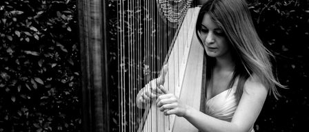 A woman dressed in a white gown plays a harp in a garden, with her hair flowing freely.