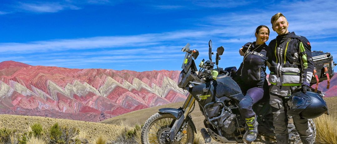 Ein Motorrad mit zwei Fahrern steht in der Wüste. Die Fahrer tragen Motorradausrüstung und Helme. Im Hintergrund befindet sich eine farbenfrohe Bergkette. Unten ist ein Veranstaltungsposter zu sehen.