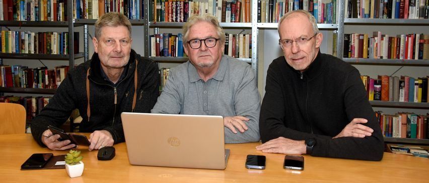 Drei Männer sitzen an einem Tisch mit Laptop, Smartphones und einer Topfpflanze. Sie sind in einem Raum mit Bücherregalen voller Bücher.