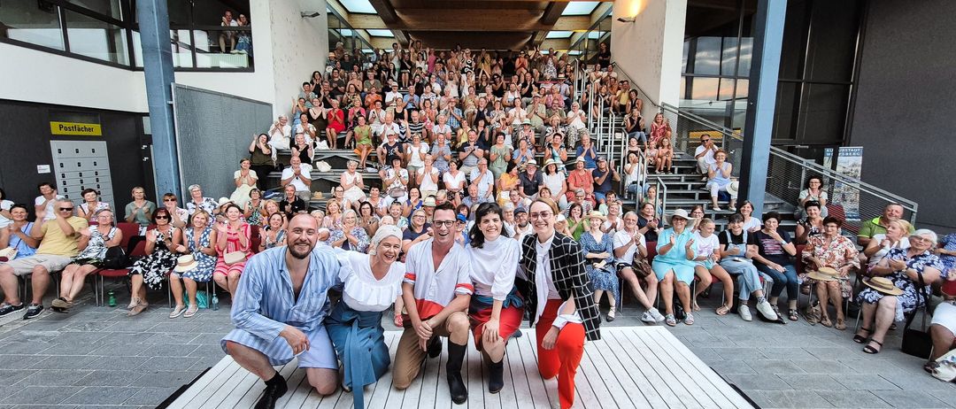 Eine Gruppe von Darstellern posiert für ein Foto auf einer Holzterrasse, mit einem Publikum, das auf den Stufen hinter ihnen sitzt, alle lächeln und klatschen.