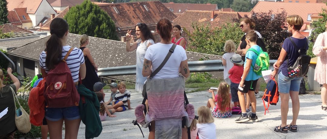 Eine Gruppe von Menschen, darunter Erwachsene und Kinder, befindet sich auf einem Dach mit Blick auf eine Stadt. Einige sitzen auf dem Boden, während andere stehen und sich unterhalten.