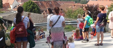 Eine Gruppe von Menschen, darunter Erwachsene und Kinder, befindet sich auf einem Dach mit Blick auf eine Stadt. Einige sitzen auf dem Boden, während andere stehen und sich unterhalten.