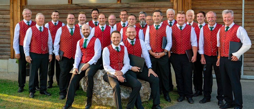 Eine Gruppe von Männern in roten Westen und grünen Krawatten posiert für ein Foto vor einem Holzgebäude.