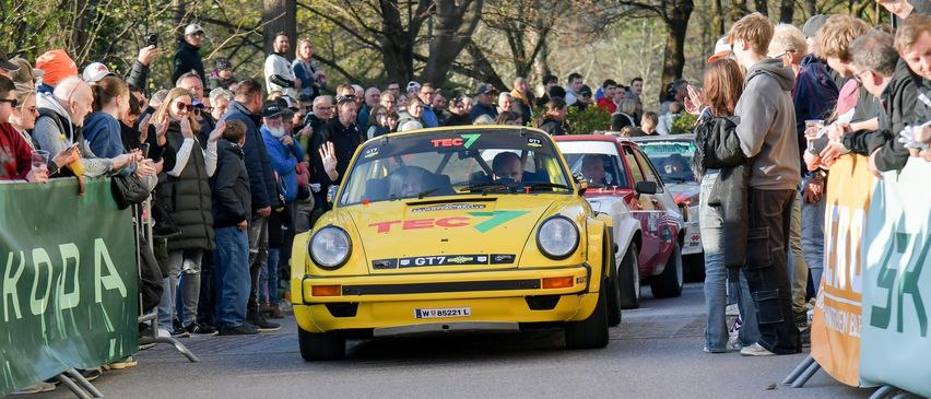 Ein gelbes Oldtimer-Auto fährt die Straße hinunter, während eine Menschenmenge zusieht. Das Auto hat die Aufschrift TEC und GT7 auf der Front. Die Menschenmenge steht am Straßenrand, und einige machen Fotos.