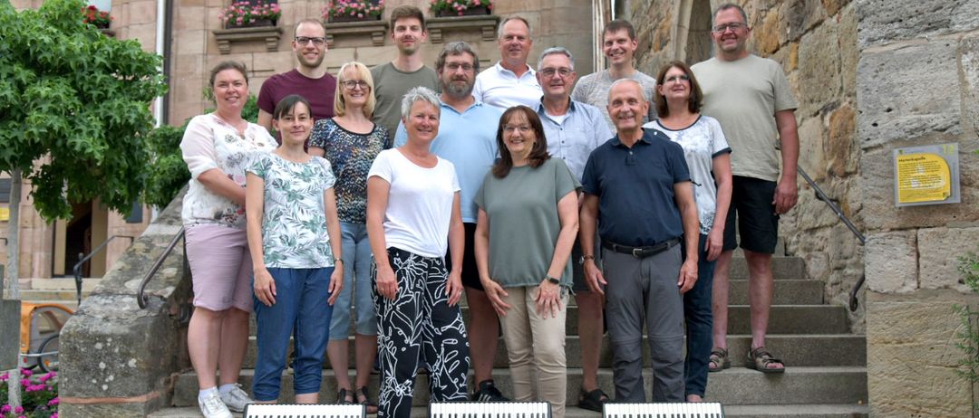 Eine Gruppe von Menschen steht auf einer Treppe, lächelt und posiert für ein Foto. Sie halten Akkordeons vor sich. Das Gebäude hinter ihnen hat eine Steinmauer und Glasfenster.