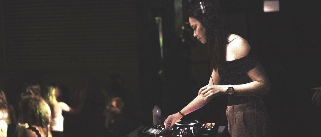 Eine Frau ist als DJ bei einer Party unterwegs, trägt Kopfhörer und eine Uhr und hat einen Plattenspieler und Gläser auf dem Tisch.