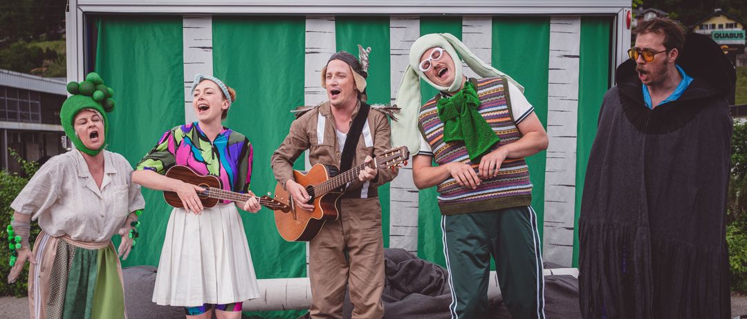 Drei Darsteller auf der Bühne, einer spielt Gitarre, ein anderer singt und ein Dritter trägt ein Hasenkostüm. Ein grüner und weißer Hintergrund ist hinter ihnen.