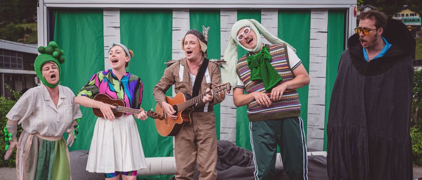 Drei Darsteller auf der Bühne, einer spielt Gitarre, ein anderer singt und ein Dritter trägt ein Hasenkostüm. Ein grüner und weißer Hintergrund ist hinter ihnen.