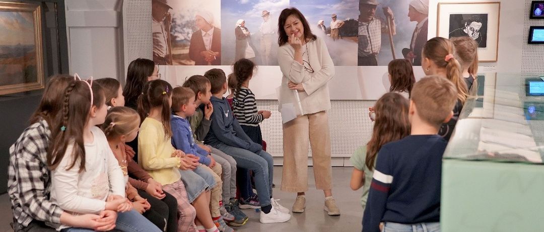 A group of children and a woman are sitting in a room, possibly in a museum. The woman is standing and holding a book. The children are sitting on a red stool. The image has a watermark with the text 'Museum im Lavanthaus'.