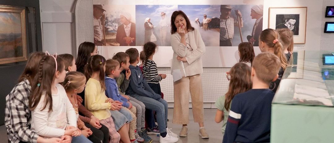 Eine Gruppe von Kindern und einer Frau sitzen auf einer Bank in einem Museum. Die Frau steht und hält ein Buch. Die Kinder schauen sich etwas an. Das Museum hat eine Glasvitrine. Der Text auf dem Plakat lautet Museum im Lavanthaus Workshops für Kinder und Jugendliche in den Sommerferien.