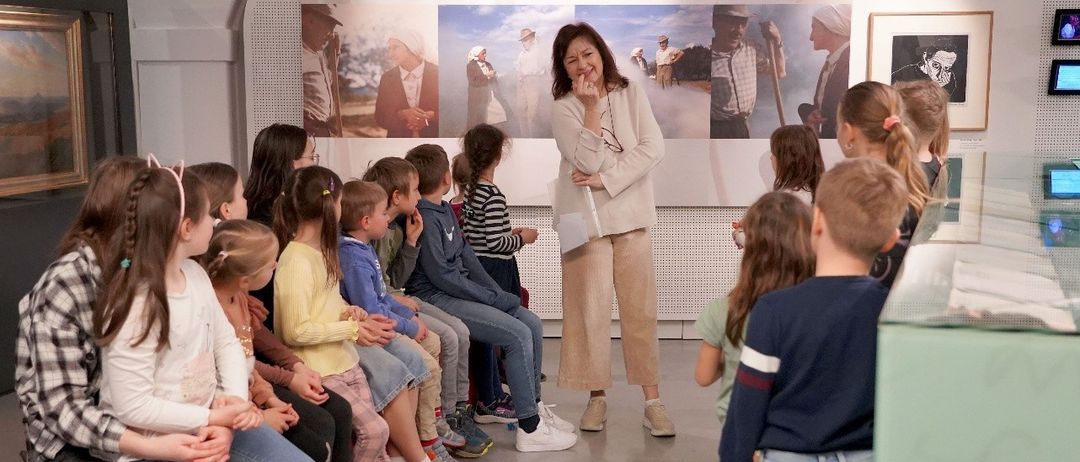 Eine Gruppe von Kindern und einer Frau sitzen in einem Raum. Eine Frau steht und spricht mit ihnen. Das Bild hat Text, der 'Museum im Lavanthaus' und 'Workshops für Kinder und Jugendliche in den Sommerferien' lautet.