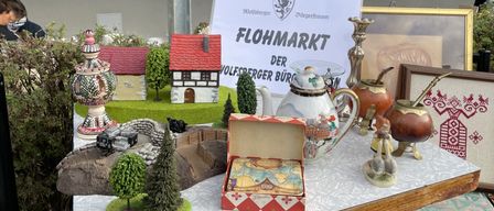 Ein Tischdisplay mit Miniaturmodellen von Häusern, einem Teepott und verschiedenen anderen Gegenständen, mit einem Schild, das 'Flohmarkt' davor steht.