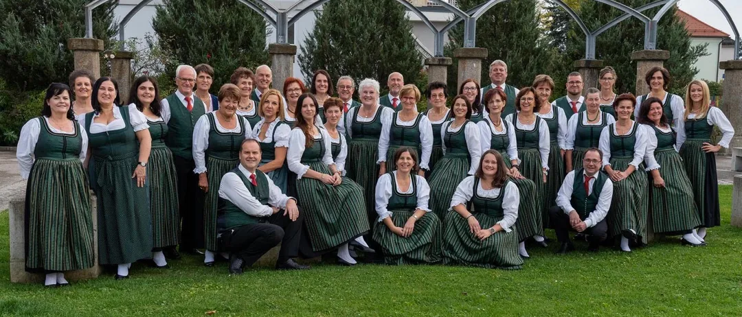 Eine Gruppe von Menschen in traditioneller grüner und weißer Kleidung, die für ein Foto vor einem Garten mit bogenförmigen Strukturen und Bäumen posiert.