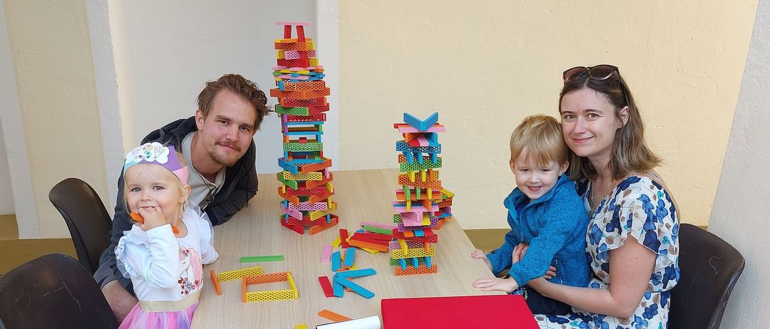 Eine vierköpfige Familie sitzt um einen Tisch mit bunten Bauklötzen. Der Mann und die Frau lächeln, und die beiden Kinder spielen mit den Bauklötzen. Der Tisch hat eine rote Matte, und in der Nähe steht ein Aufbewahrungsbehälter.