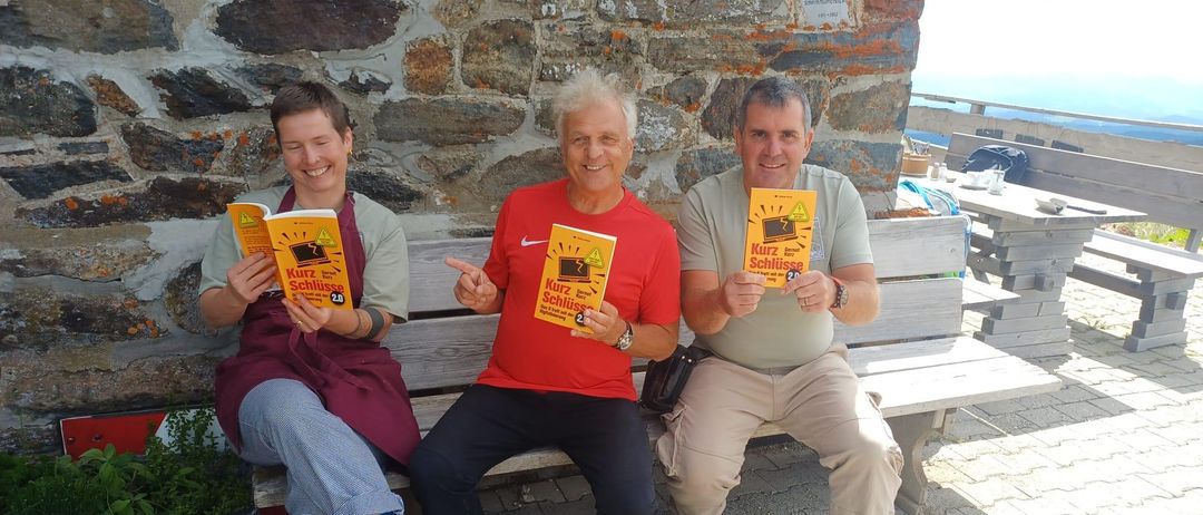 Drei Männer sitzen auf einer Bank, jeder hält ein Buch mit dem Titel 'Kurz Schliesse'. Sie lächeln und scheinen für ein Foto zu posieren. Hinter ihnen befindet sich eine Steinmauer mit einem Speisezettel.