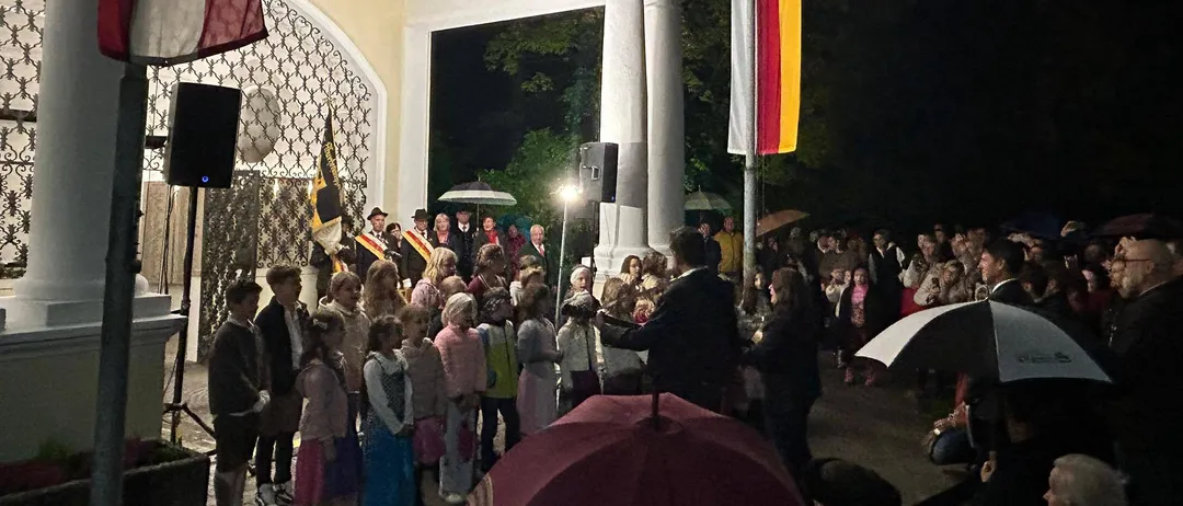 Eine nächtliche Veranstaltung mit einer Menschenmenge, einige mit Regenschirmen, vor einem Gebäude mit Fahnen und einer Bühne.