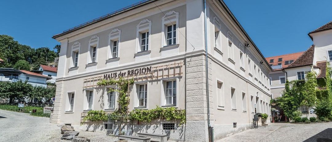 Ein großes weißes Gebäude mit mehreren Fenstern und einem Schild, das 'Haus der Region' besagt. Das Gebäude ist von einem Kopfsteinpflasterweg umgeben und hat einige Steine und Pflanzen davor.