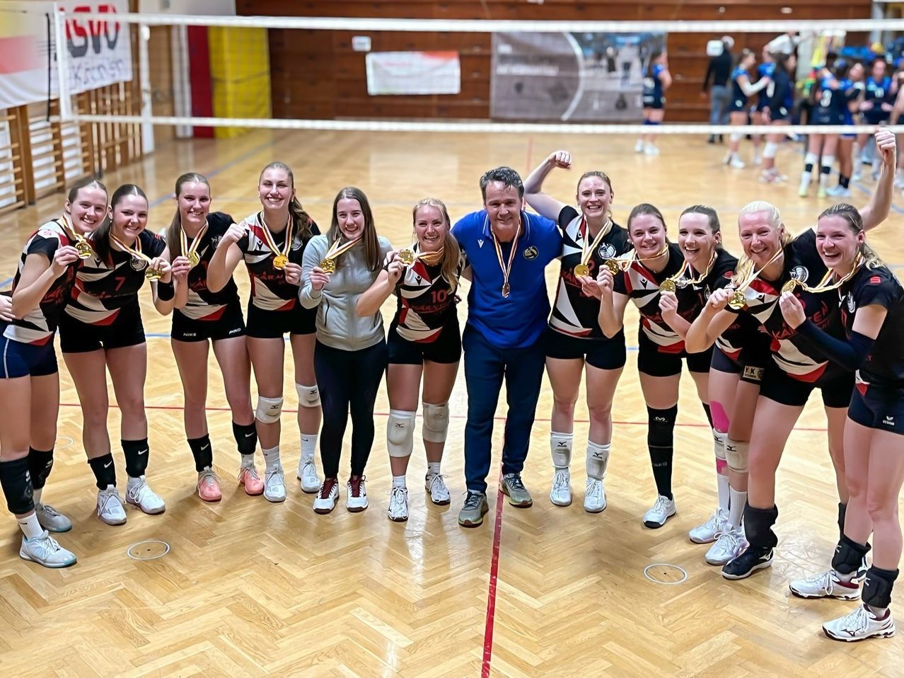 Eine Gruppe junger Volleyballspielerinnen posiert mit ihrem Trainer für ein Foto. Sie tragen alle Medaillen und lächeln für die Kamera.