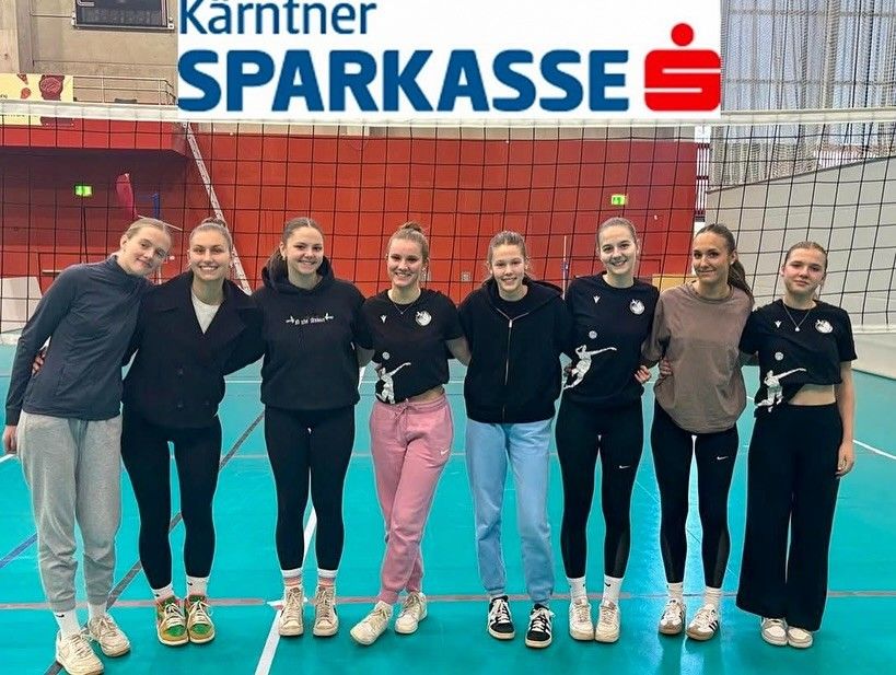 Eine Gruppe von Frauen steht in einer Reihe auf einem Volleyballplatz, trägt passende Jacken mit dem Logo 'Karntner Sparkasse' und lächelt für ein Foto.
