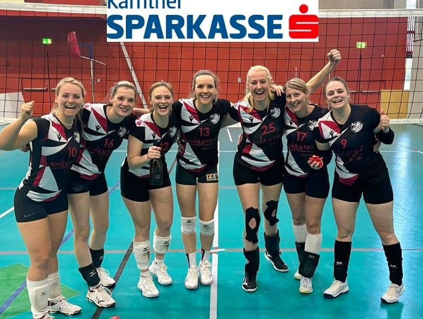Sieben Frauen stehen in einer Reihe auf einem Volleyball-Spielfeld, lächeln und posieren für ein Foto. Sie tragen alle passende Uniformen mit Nummern auf dem Rücken.