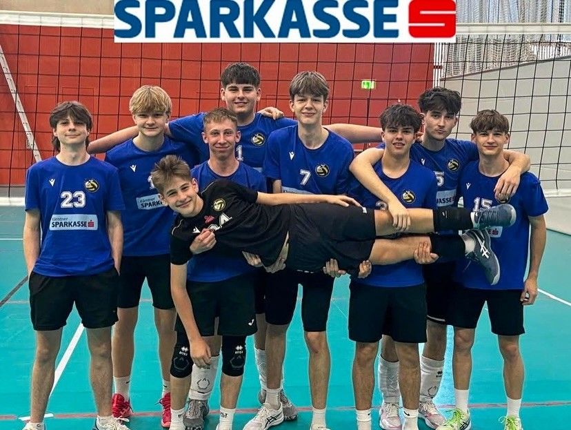 Eine Gruppe junger männlicher Athleten in blauen Uniformen und Turnschuhen posiert für ein Foto auf einem Volleyballplatz. Einer von ihnen liegt auf dem Rücken, wobei seine Beine von seinen Teamkollegen gestützt werden.