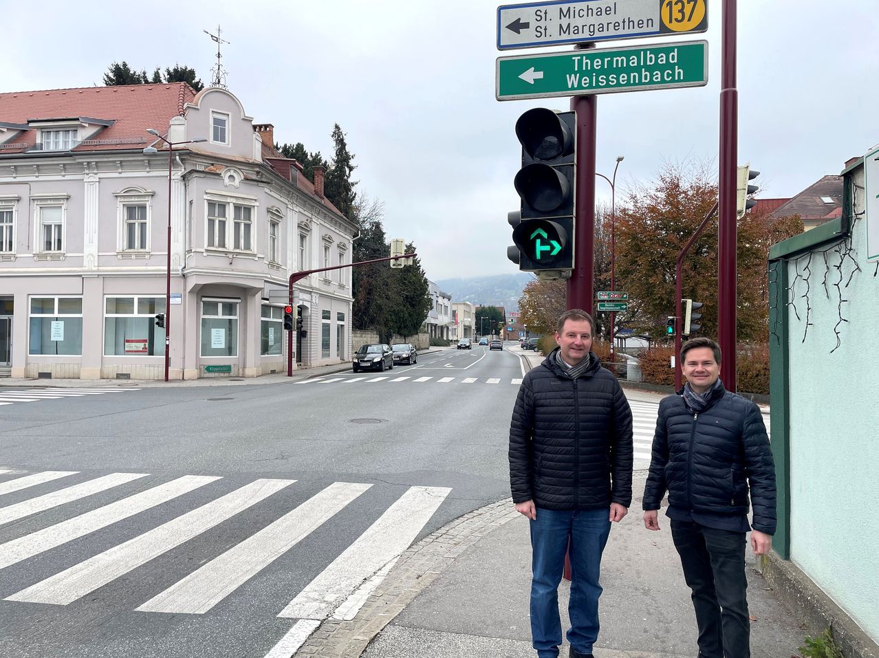 Zwei Männer stehen an einer Straßenecke neben einer Ampel. Sie lächeln und schauen in die Kamera. Straßenschilder zeigen Richtungen nach St. Michael, St. Margarethen und Thermalbad Weissenbach.