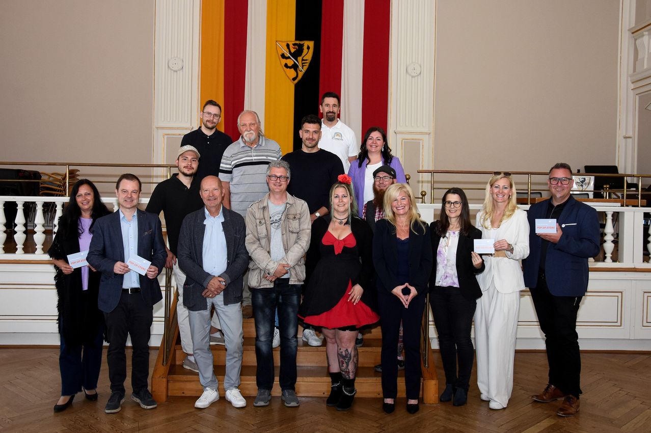 Gruppe von Menschen in unterschiedlicher Kleidung und Schuhen, die vor einer deutschen Flagge und einem Wappen für ein Foto posieren.
