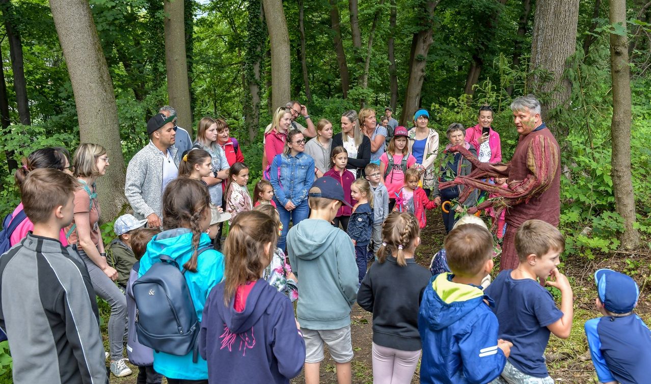 Eine Gruppe von Menschen, einschließlich Kindern, versammelt sich in einem Wald. Sie hören einem Mann zu, der eine Pflanze in der Hand hält. Sie alle schauen zu dem Mann. Einige von ihnen machen Fotos mit ihren Handys. Das Gebiet ist von Bäumen umgeben.