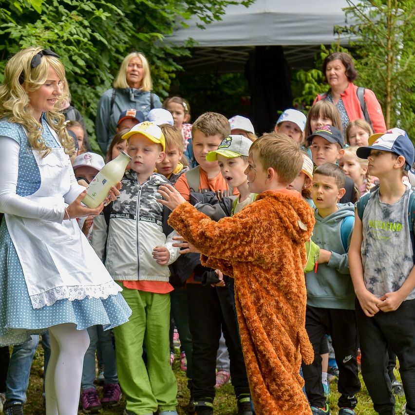 Eine Frau in einem weißen Kleid hält eine Flasche und spricht zu einer Gruppe von Kindern, einige in Kostümen, stehend auf Gras mit Bäumen im Hintergrund.