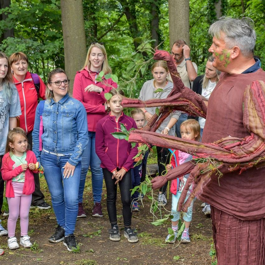 Eine Gruppe von Erwachsenen und Kindern in einem Wald, ein Mann hält ein großes rotes Objekt, das einer Spinne ähnelt, und eine Frau in einer Jeansjacke lächelt.