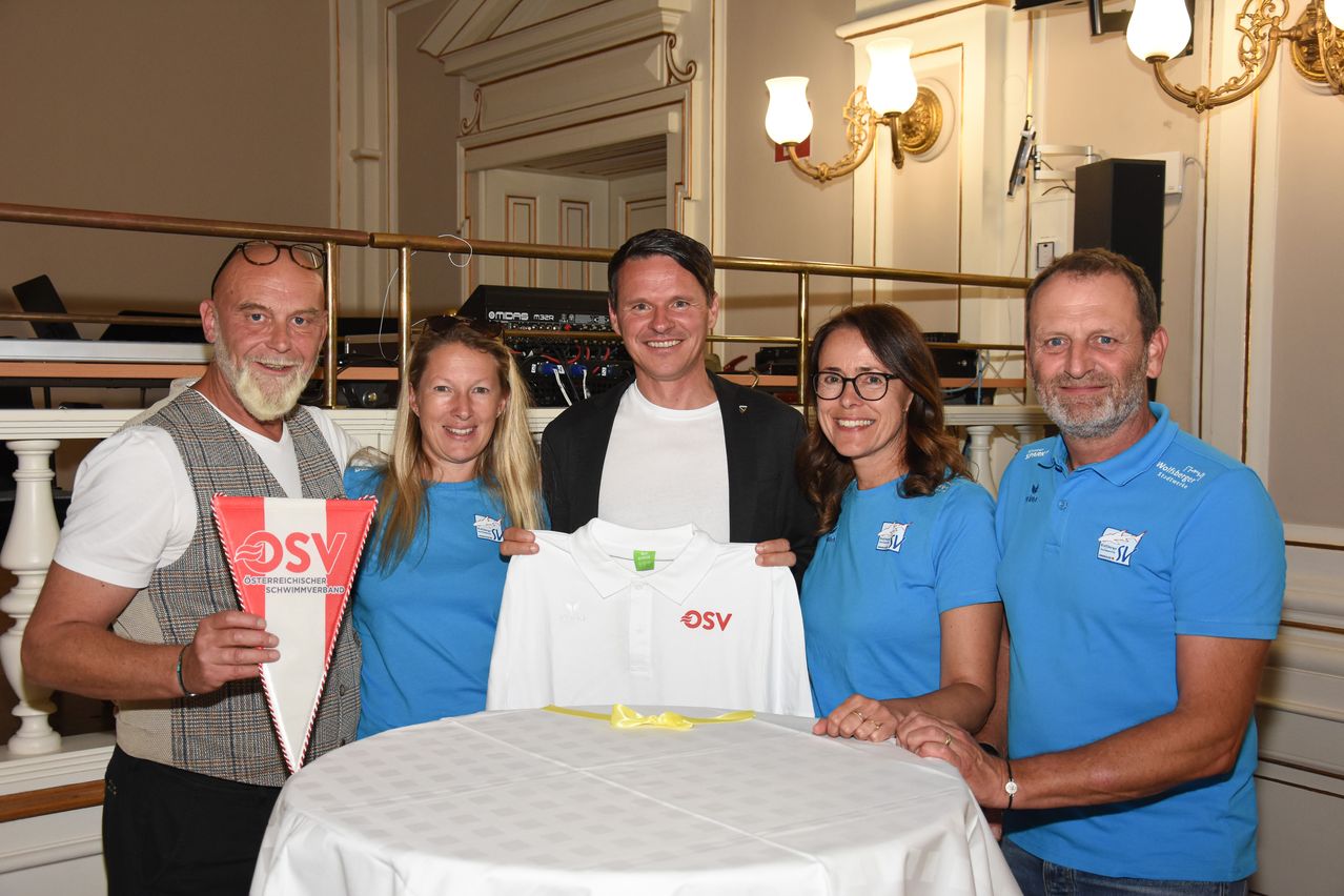 Fünf Personen posieren für ein Foto mit einem Tisch und einem weißen Shirt. Ein Mann hält eine Flagge. Eine Frau hält ein weißes Shirt. Ein Mann lächelt. Eine weitere Frau trägt eine Brille. Ein weiterer Mann lächelt.