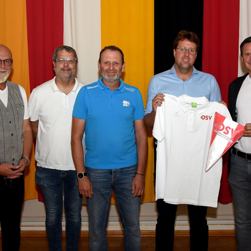 Fünf Männer stehen vor einem Hintergrund mit den deutschen Flaggenfarben. Einer hält ein weißes Shirt mit Logo und Etikett.