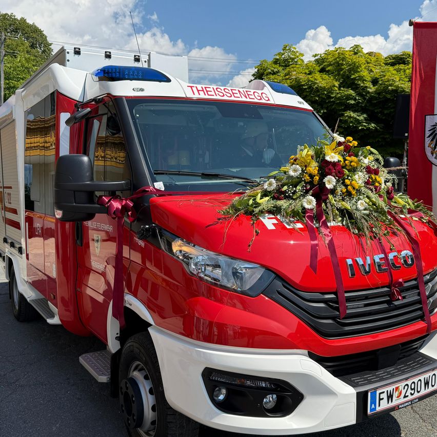 Ein roter und weißer Iveco-Feuerwehrwagen, geschmückt mit Blumen, steht auf einem Freigelände. Rechts ist eine rote Flagge zu sehen und der Himmel ist klar mit einigen Wolken.