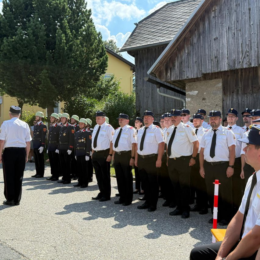 Eine Reihe von Feuerwehrleuten in Uniform steht vor einem Gebäude. Eine Person sitzt auf einer Bank vor ihnen.