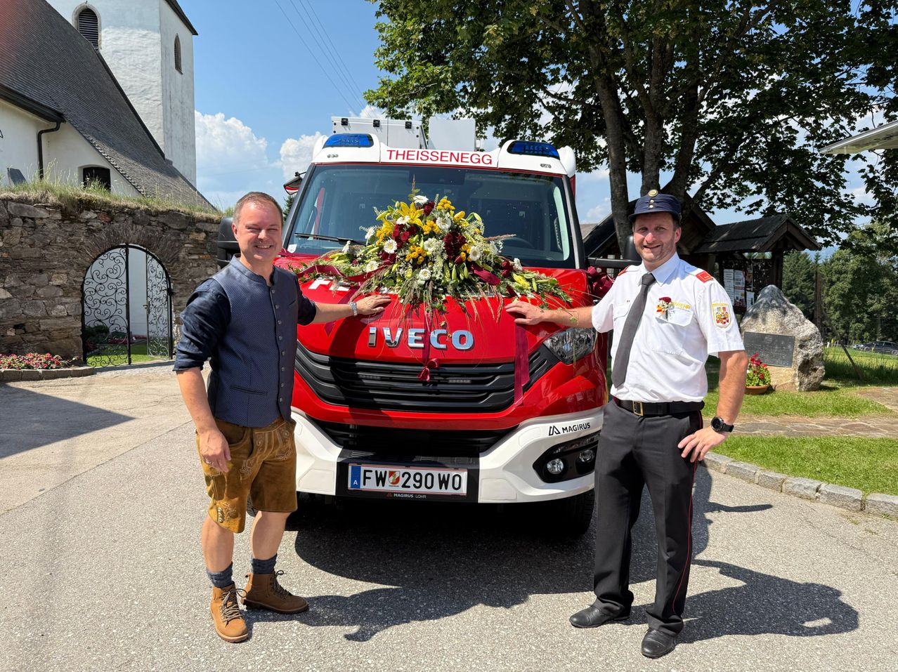 Zwei Männer stehen vor einem roten Iveco-Feuerwehrfahrzeug, geschmückt mit einer Blumendekoration. Sie tragen Uniformen, einer mit einer Uhr, stehen auf einer Betonfläche.