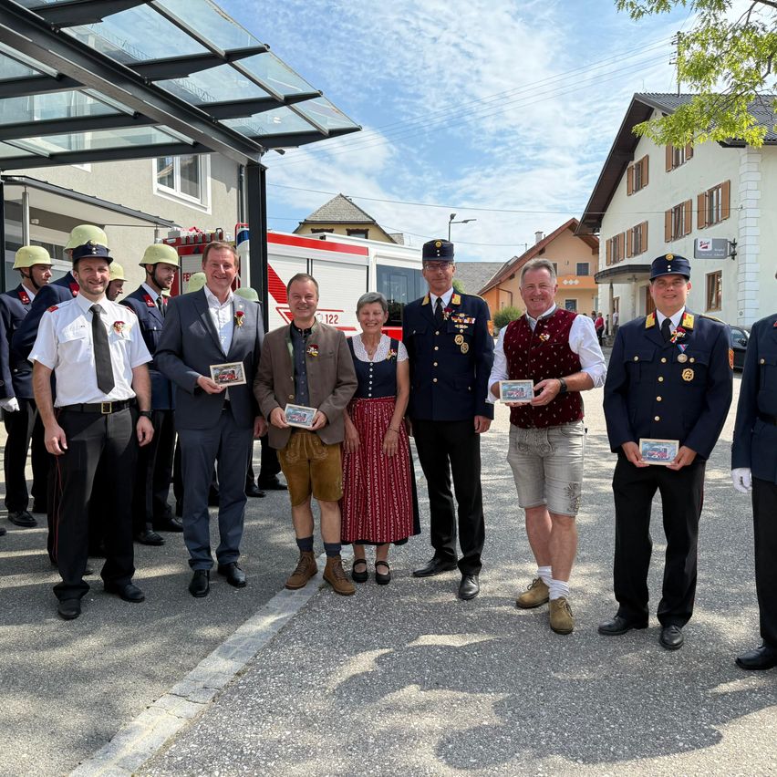 Eine Gruppe von Menschen in Uniformen, wahrscheinlich Feuerwehrleute, posiert für ein Foto vor einer Feuerwache an einem sonnigen Tag. Sie halten Auszeichnungen.