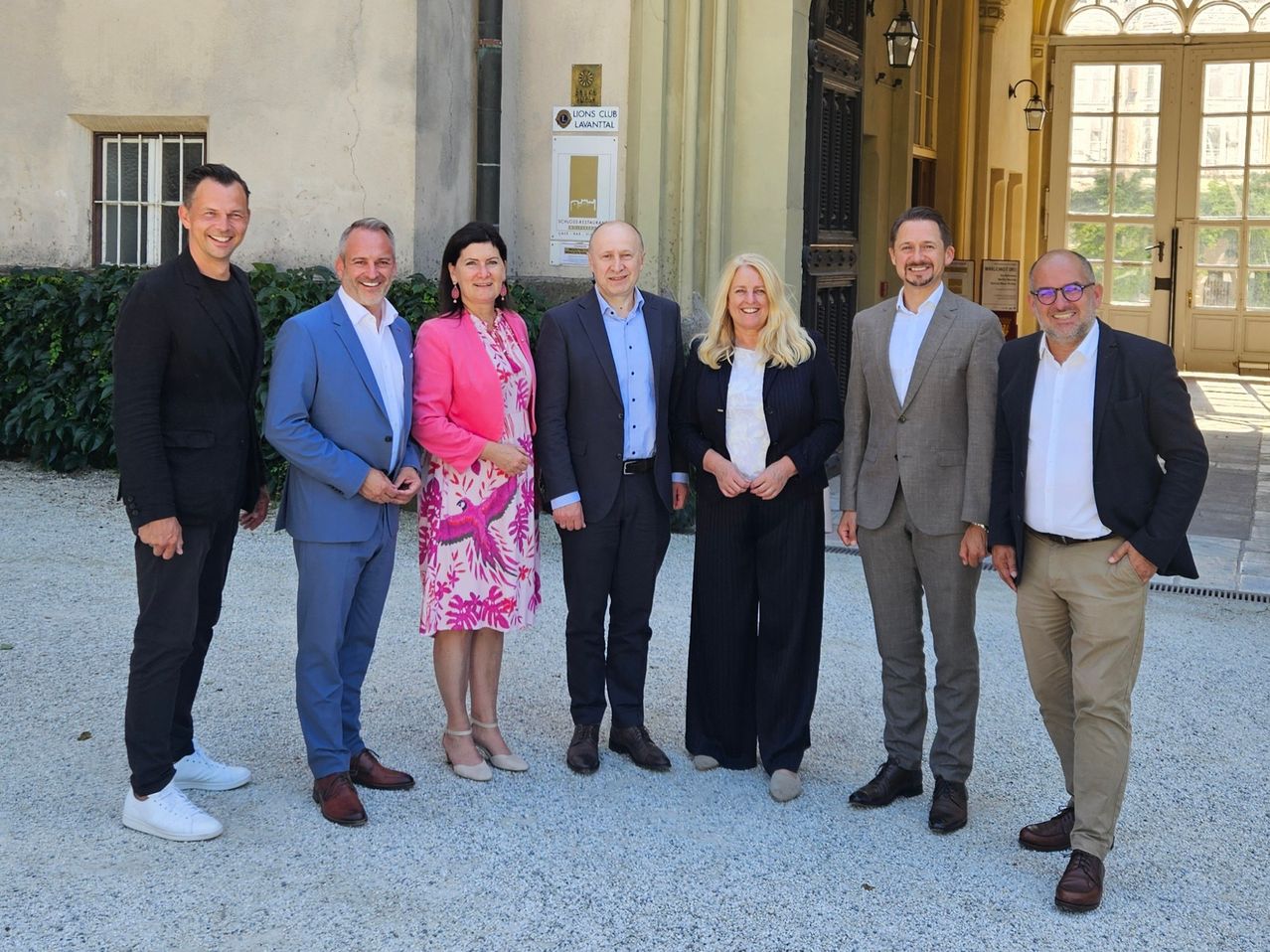 Eine Gruppe von sieben Erwachsenen steht vor einem Gebäude. Sie sind in formeller Kleidung gekleidet. Das Gebäude hat ein Schild mit der Aufschrift Lions Club Lavantaital.