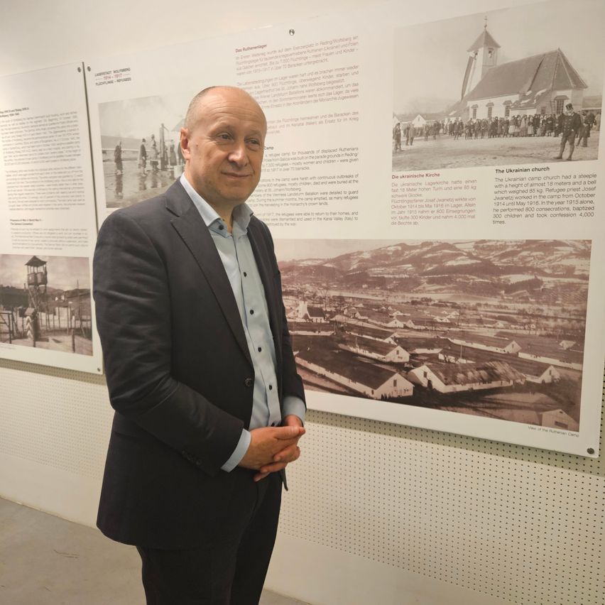 Ein Mann steht vor einem großen Wandbild und lächelt in die Kamera. Das Wandbild enthält historische Bilder und Texte, eines zeigt eine Kirche und das andere ein Lager.