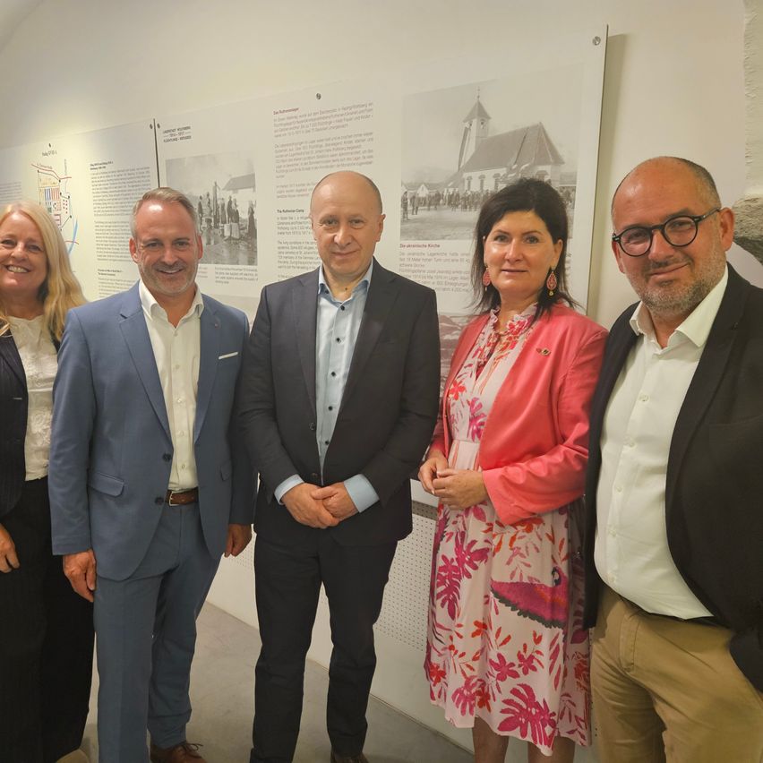 Fünf Personen stehen nebeneinander vor einer Wand mit historischen Informationen. Sie sind alle formell gekleidet.